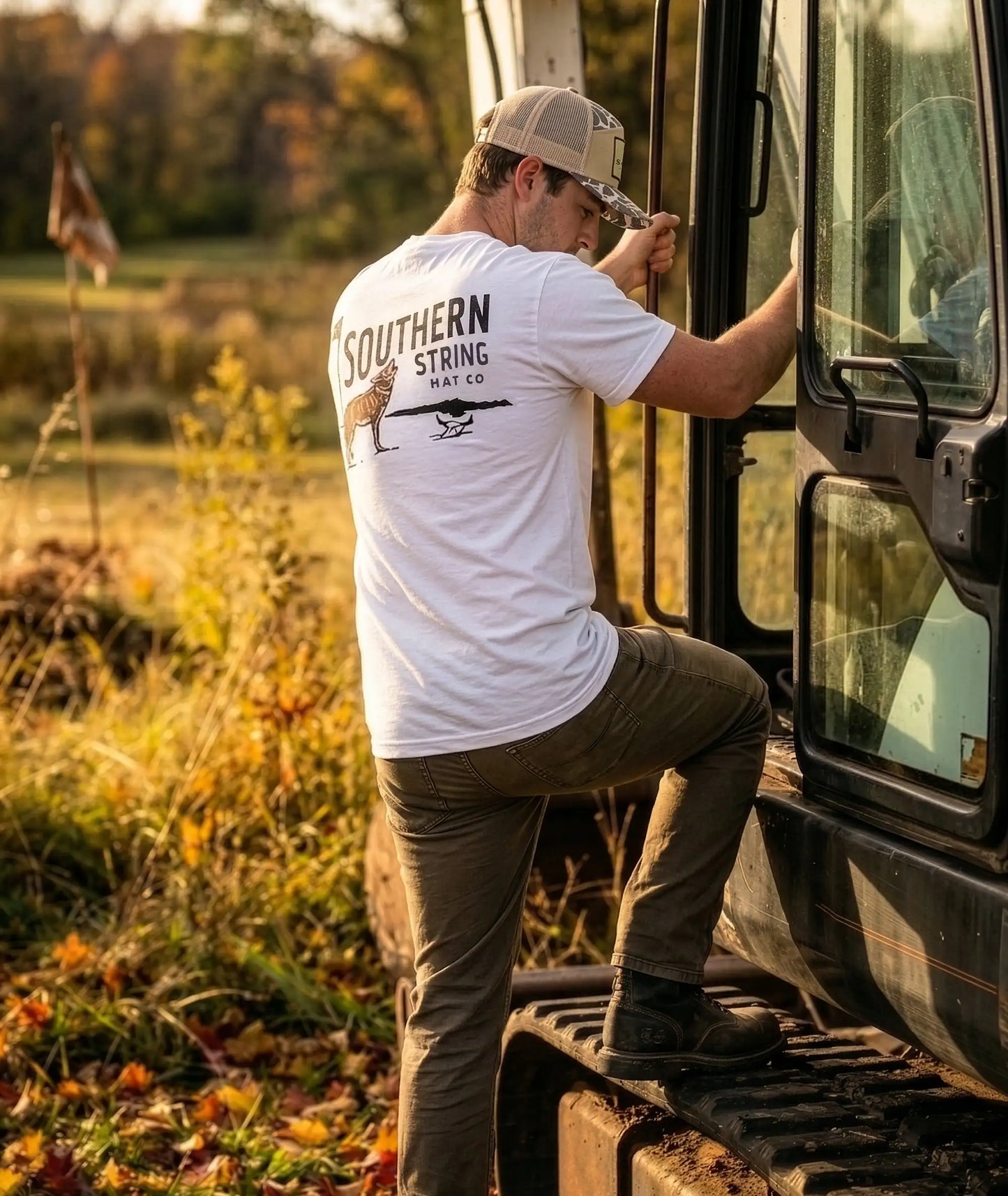 A lifestyle portrait of a male model stepping up on an excavator wearing Southern String Hat Co's 'The Yote' t-shirt featuring a coyote and golf pin illustration on a premium white colored cotton tee