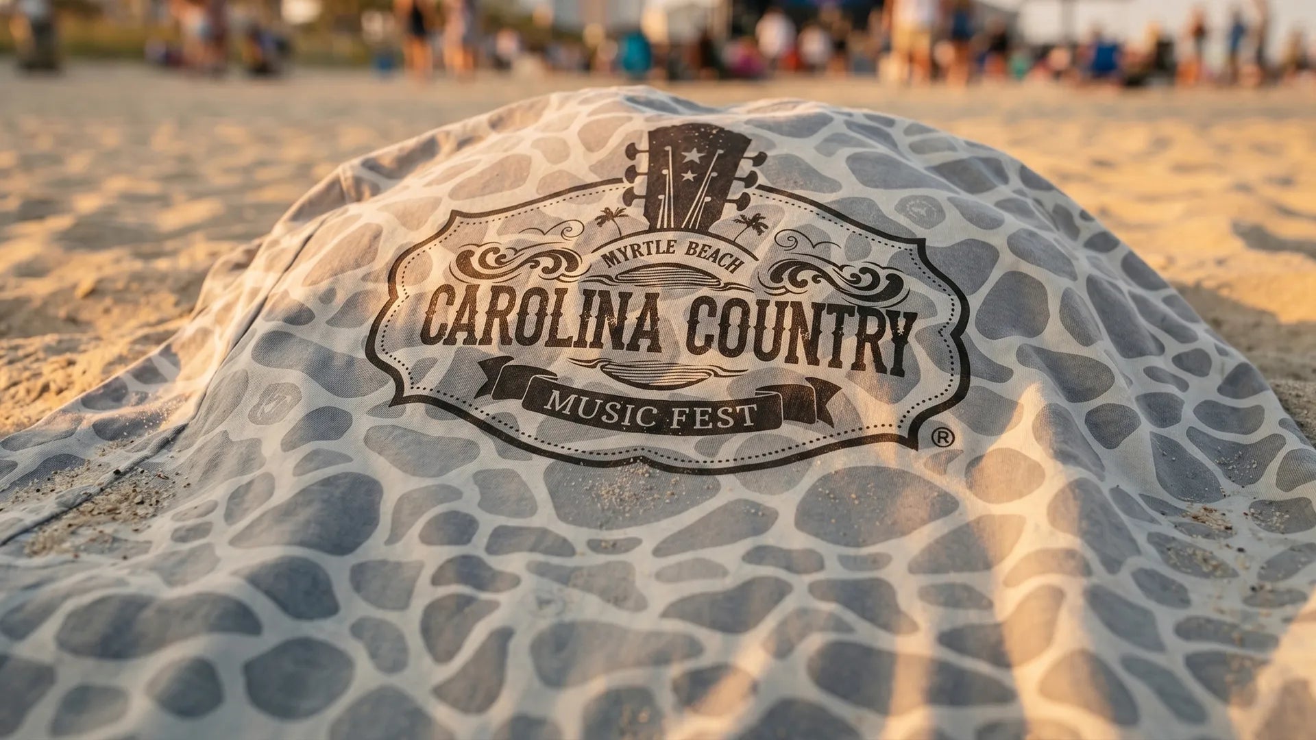 A close-up shot of the back print of the special-edition 2026 SS Hat Co x Carolina Country Music Fest camo t-shirt resting on a sandy beach at a music venue.